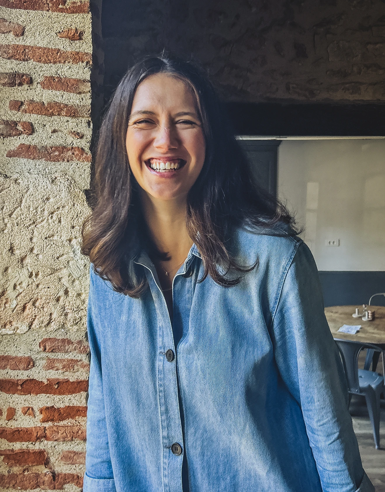 Holly leans against a brick wall, wearing a long denim shirt she smiles for the camera