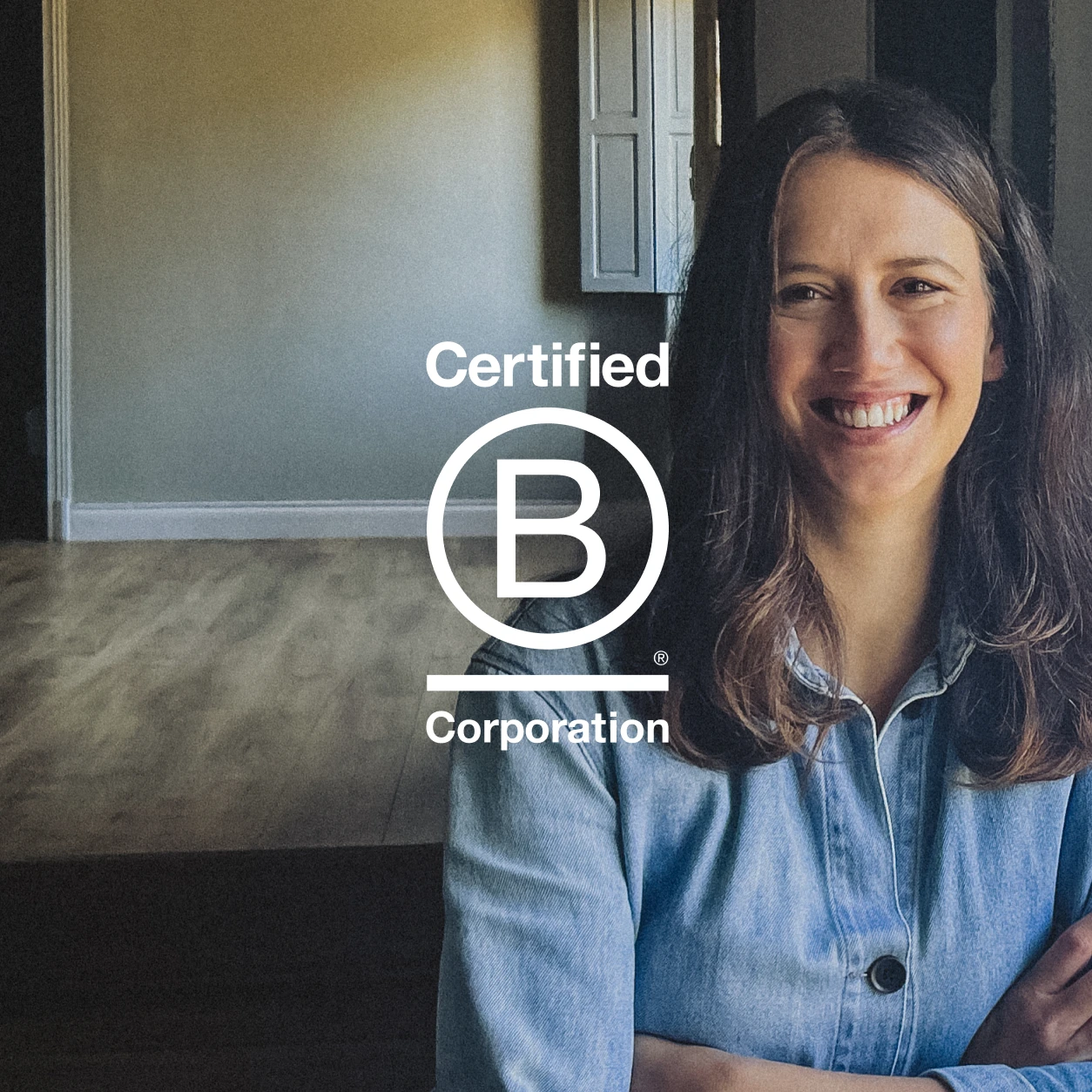Holly is smiling whilst leaning against a wall, she is wearing a denim shirt and has long brown hair. The B Corp logo is overlaid.