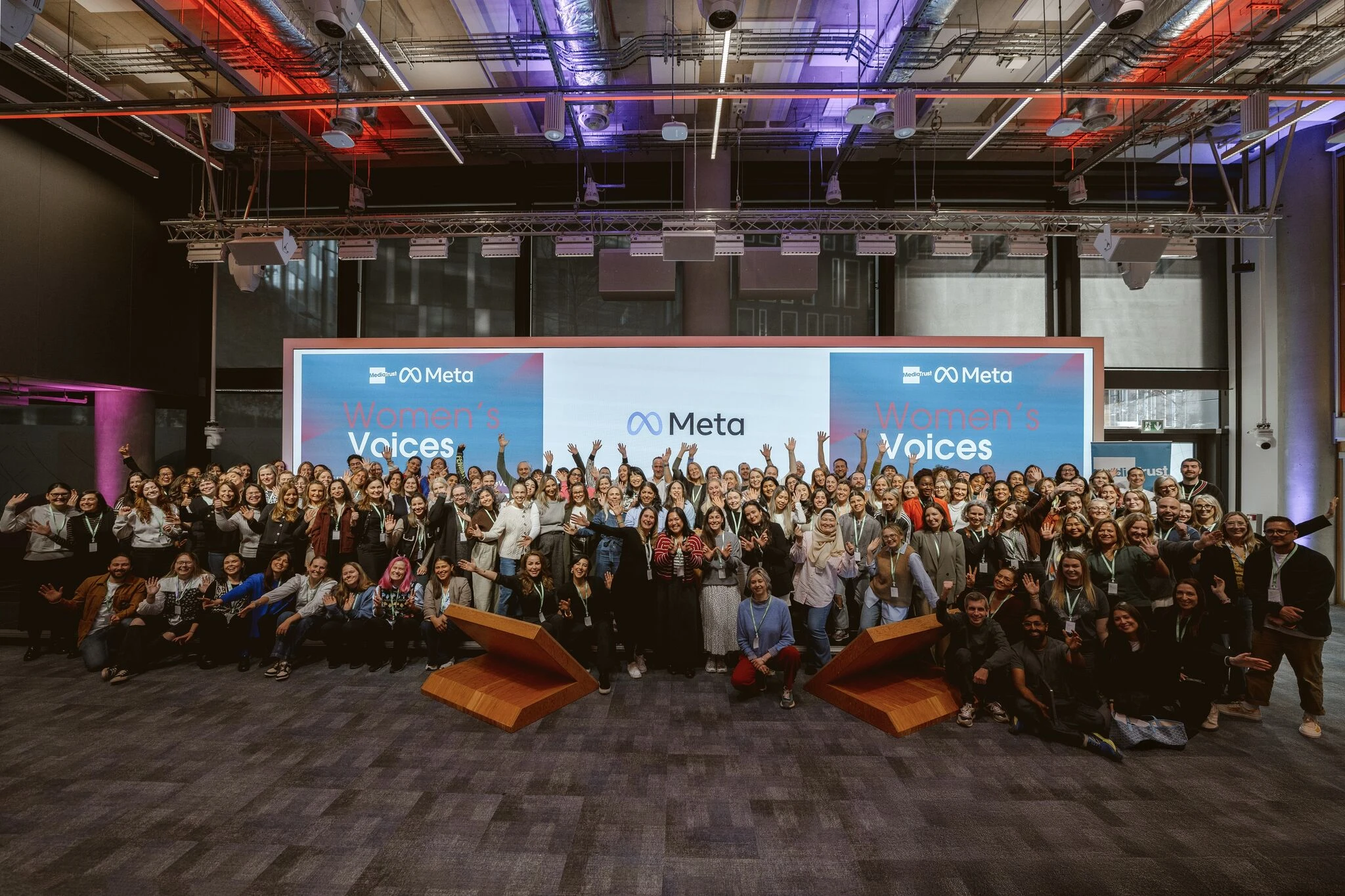 The entire group of charities and volunteers as part of Women's Voices 2025 are photographed smiling and waving in a large conference hall at the Meta offices in London