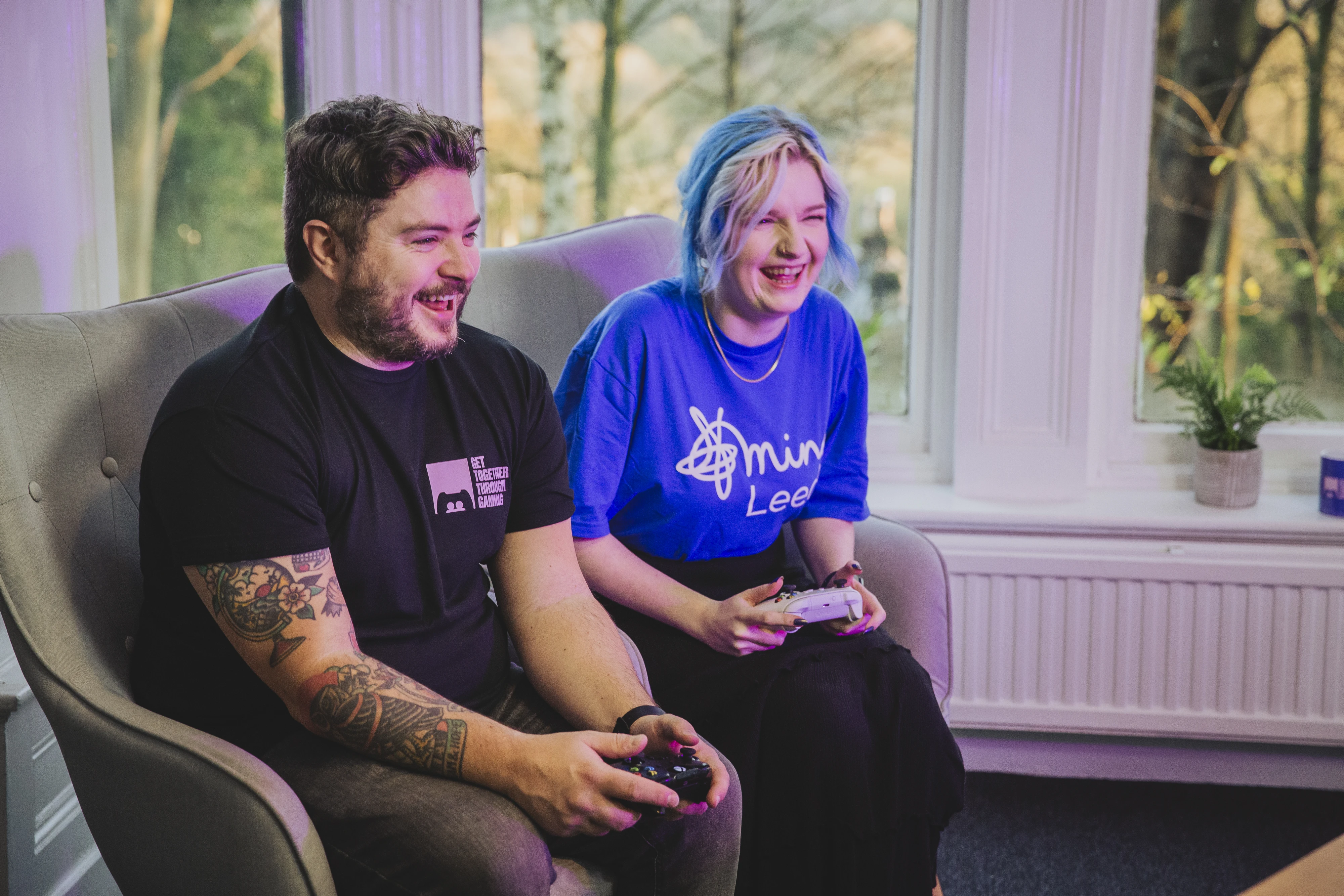 Col and Cat laugh whilst playing video games for Leeds Mind's Get Together Through Gaming, Col wears a black GTTG t-shirt, Cat wears a blue Leeds Mind t-shirt